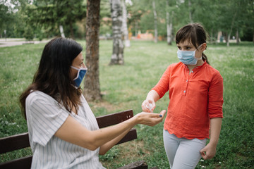 Fototapeta premium Little girl with mask holding disinfectant over woman's hands outdoor. Cute girl with antivirus mask holding hand sanitizer while standing in park near her mom who is sitting on bench.