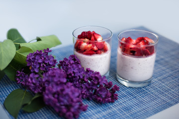 Layered strawberry homemade italian creamy dessert panna cotta with fresh berries in glass on blue background with branch of purple lilac. Organic sweet. Side view of strawberry panna-cotta