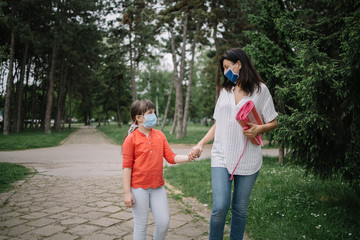 Woman and child holding their hands while walking in park. Sad little girl with antivirus mask looking at her mother while holding hands and walking in city park.