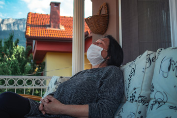 Middle aged caucasian woman with medical face mask, resting on cpuch on veranda during COVID-19 threat. Woman with mask on her terrace is resting, sleeping, looking, talking.