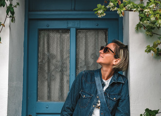 Pretty fashionable woman standing in doorway of old house. Cool dressed lady in front of turquoise wooden door with flowers around.