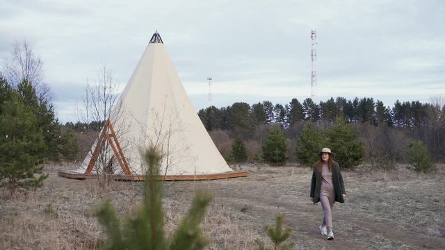 Young nice woman in hat walking through wooded countryside to glamping outside teepee / tent / yurt at sunset. ecotourism, forest
