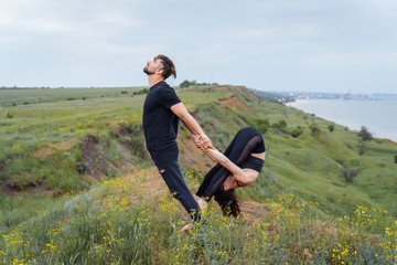 Naklejka na ściany i meble Young caucasian couple practicing acro yoga together on the hill near the sea. Couple, partner yoga. Athlete flexing beautifully and holding girl with his trained hands. Sportswoman stretching