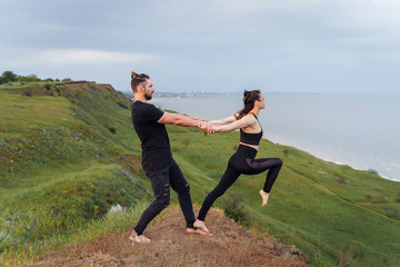 Handsome, attractive athlete in a black T-shirt and jeans holding the hand of his athletic girl. Doing acroyoga in the air outdoors. Sportswoman making step into the abyss from the cliff. Partner yoga