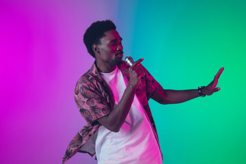 African-american male singer portrait isolated on gradient studio background in neon light. Beautiful male model with microphone. Concept of human emotions, facial expression, ad, music, art.