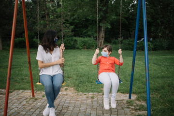 Woman and little girl on swing while wearing antivirus masks. Mother and daughter with protection masks are swinging in park.