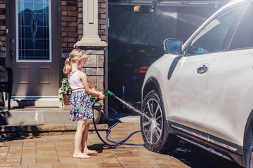 Young preschool girl washing car on driveway in front house on summer day. Kids home errands duty chores responsibilities concept. Child playing with a hose spraying water. Home work lifestyle. - obrazy, fototapety, plakaty