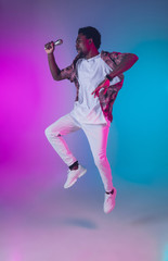 Fototapeta na wymiar African-american male singer portrait isolated on gradient studio background in neon light. Beautiful male model with microphone. Concept of human emotions, facial expression, ad, music, art.