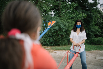 Mother and daughter playing with a rocking chair in the park. Back of a young girl wearing mask in the foreground, playing with her mother, also using protection mask, enjoying free time in the park.