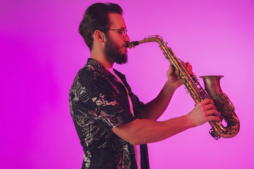 Young caucasian jazz musician playing the saxophone on gradient pink studio background in neon light. Concept of music, hobby, festival. Joyful, cheerful attractive guy. Colorful portrait of artist.