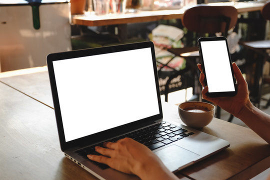 Mockup image of a woman holding blank mobile phone while using laptop with blank  screen on table in cafe.