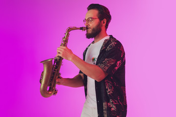 Young caucasian jazz musician playing the saxophone on gradient pink studio background in neon...