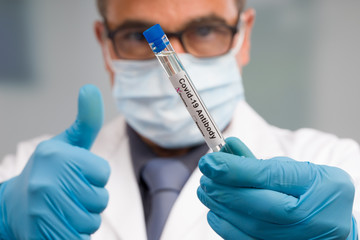 Scientist or doctor with medical face mask and medical gloves is handling virus test tube with antibody result markers in front of laboratory in background and shows thumbs up