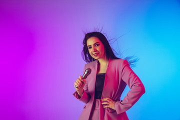Naklejka premium Caucasian female singer portrait isolated on gradient studio background in neon light. Beautiful female model in pink wear with microphone. Concept of human emotions, facial expression, ad, music, art