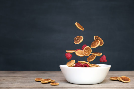 Flying tiny pancake cereal - trendy quarantine food. Mini pancakes and fresh raspberries in motion falling in white bowl on black chalkboard background. Levitation, gravity. Homemade breakfast.
