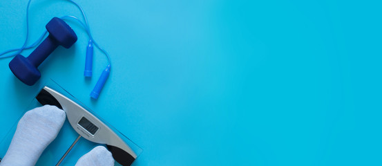 Woman measures her weight on scale on a blue background with skipping rope and dumbbells. Healthy lifestyle banner