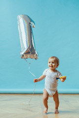 Funny kid running happy wearing pampers with one year balloon on blue background. First birthday celebration copy space