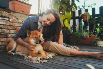 Young beautiful and positive woman hugging her dog while enjoying sunny day on home terrace with plants, Friendship Happiness, Millennial hipster girl in casual look laughing playing with pet at home