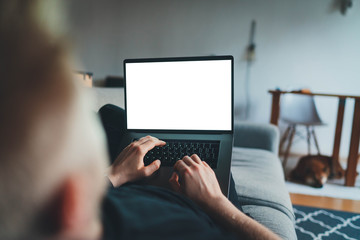 Young man relaxing on sofa at home working on laptop with blank screen, hipster guy shopping online or having video call while chilling at home on couch his dog in front of him, mockup of laptop