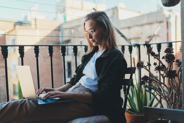 Young talented creative designer working from home on laptop computer sitting on the balcony enjoying sunny day, Millennial hipster girl an advertising copywriter in trendy look typing on keyboard