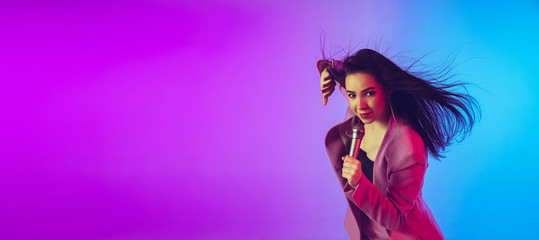 Caucasian female singer portrait isolated on gradient studio background in neon light. Beautiful female model in pink wear with microphone. Concept of human emotions, facial expression, ad, music, art