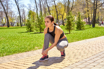 Attractive young sports woman sitting on knee and tying shoe laces on sneakers while having rest during morning jog outdoors. Beautiful fitness girl in stylish sportswear ties shoelace on sports shoes
