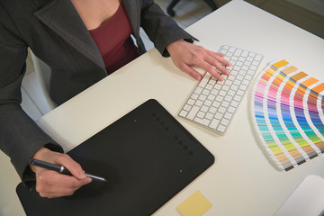 Woman freelance graphic designer at work in her home office chooses colors from palette for design and printing a new job. She work on a white desk with the black graphic tablet and computer keyboard.