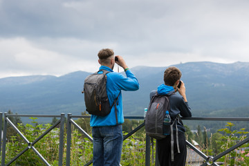 Mężczyzna i nastoletni chłopak  z plecakami oglądają przez lornetki odległy szczyt górski - obrazy, fototapety, plakaty