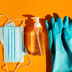 Personal protection kit when leaving the house during a coronavirus pandemic: individual face masks, gel sanitizer and hand gloves. Orange background, trend light, contrasting shadows. Top view.
