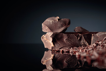 Pieces of dark bitter chocolate and chocolate chips on a black reflective background.