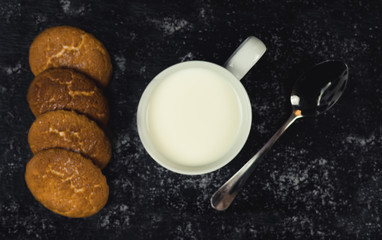Taza llena de leche blanca con montón de galletas doradas espolvoreado con azúcar y una cuchara metálica sobre una tabla de pizarra