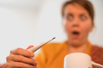 Caucasian surprised young girl checks the temperature with a thermometer. The woman has fever and headache. Disease development concept.