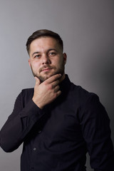 Fototapeta premium Young Bearded Dark Haired Man In Black Stylish Shirt On Gray Background, Portrait Of Confident, Charismatic Male Looking Straight, He Puts A Hand To His Chin. Close Up