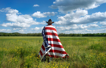 Man wearing cowboy hat standing in grassy agricultural field with American flag draped over his shoulders, blue sky white clouds