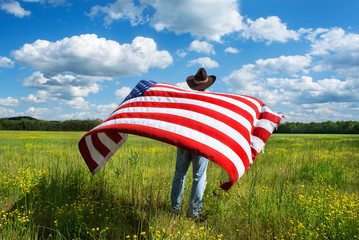 Man wearing cowboy hat standing in grassy agricultural field with American flag blowing in wind,...