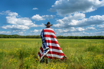 Man wearing cowboy hat with American flag draped around him standing in green agricultural field,...