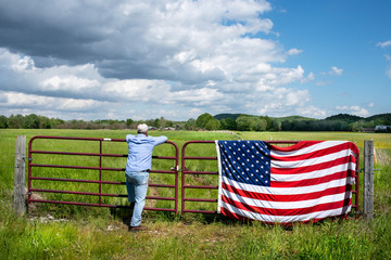 Farmer leaning on metal fence in grassy agricultural field, American flag draped over fence, blue...