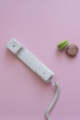 A close up view of A white vintage dial telephone handset and two macaronis on pink background. Communication device. A retro receiver with phone curly wire and copy space. Call center. Office context