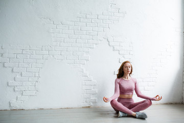 Remote plan. Red haired yoga trainer in pink tracksuit sits in lotus position with hands on knees in the white studio