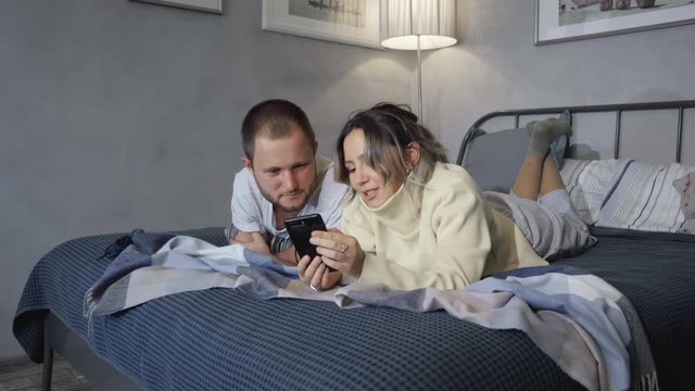 Young couple in hotel bed read texts on smartphone and laugh