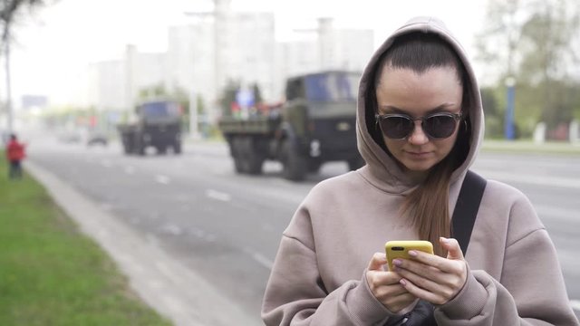 girl is typing on the phone. military vehicles are driving in the background. medium close up