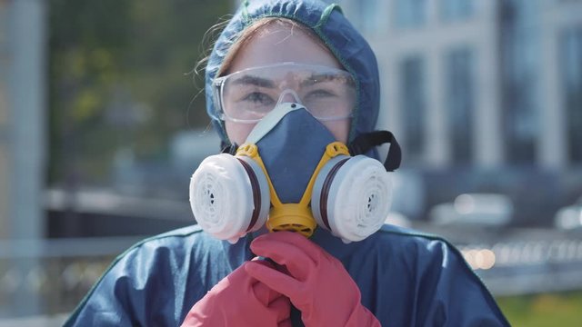 Close-up portrait of young beautiful Caucasian woman in chemical suit and protective eyeglasses putting on respirator and looking at camera. Confident female specialist posing outdoors on summer day.