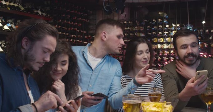 Caucasian male football fans with girlfriends watching match in pub, sending messages from mobile phones. Guys placing bets on sport game result. Men raising rates of bookmakers by cellphones in bar.