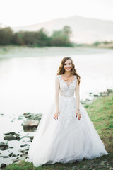 Fototapeta na wymiar Beautiful brunette bride in elegant white dress posing near river