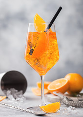 Glass of aperol spritz summer cocktail with oranges and strainer with bar spoon and cocktail shaker on light background. Ice cubes and black straw.