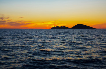 Colorful sunset and cloudy sky over the dark rocky islands against the dark blue sea.