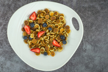 Pancake cereal. Trendy food, mini cereal pancakes in bowl on the table, grey cement background. Trendy baked mini pancakes with strawberries, blueberries . Copy space. Flat layout. Table top view.