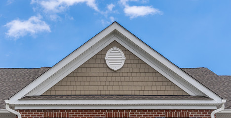 Beige shake and shingle covered gable roof with white oval attic window on a new American home with blue sky background
