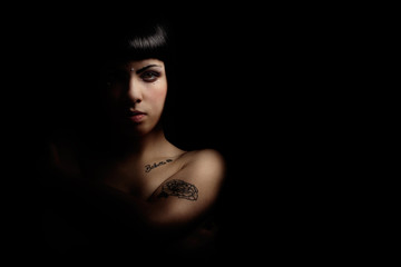 Mystery beautiful young woman in shadow and light. A beautiful young girl is illuminated sideways to half by a dim light. On a black background. Bob and some tattoos on the body.