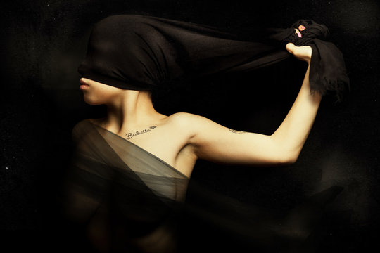 Woman Blinded By A Cloth Hiding Face. Rebellion And Silence. A Young Woman Is Blinded By A Cloth Pulled From Hand. Artistic Portrait With Black Background. A Silk Veil Covering Her Breasts.